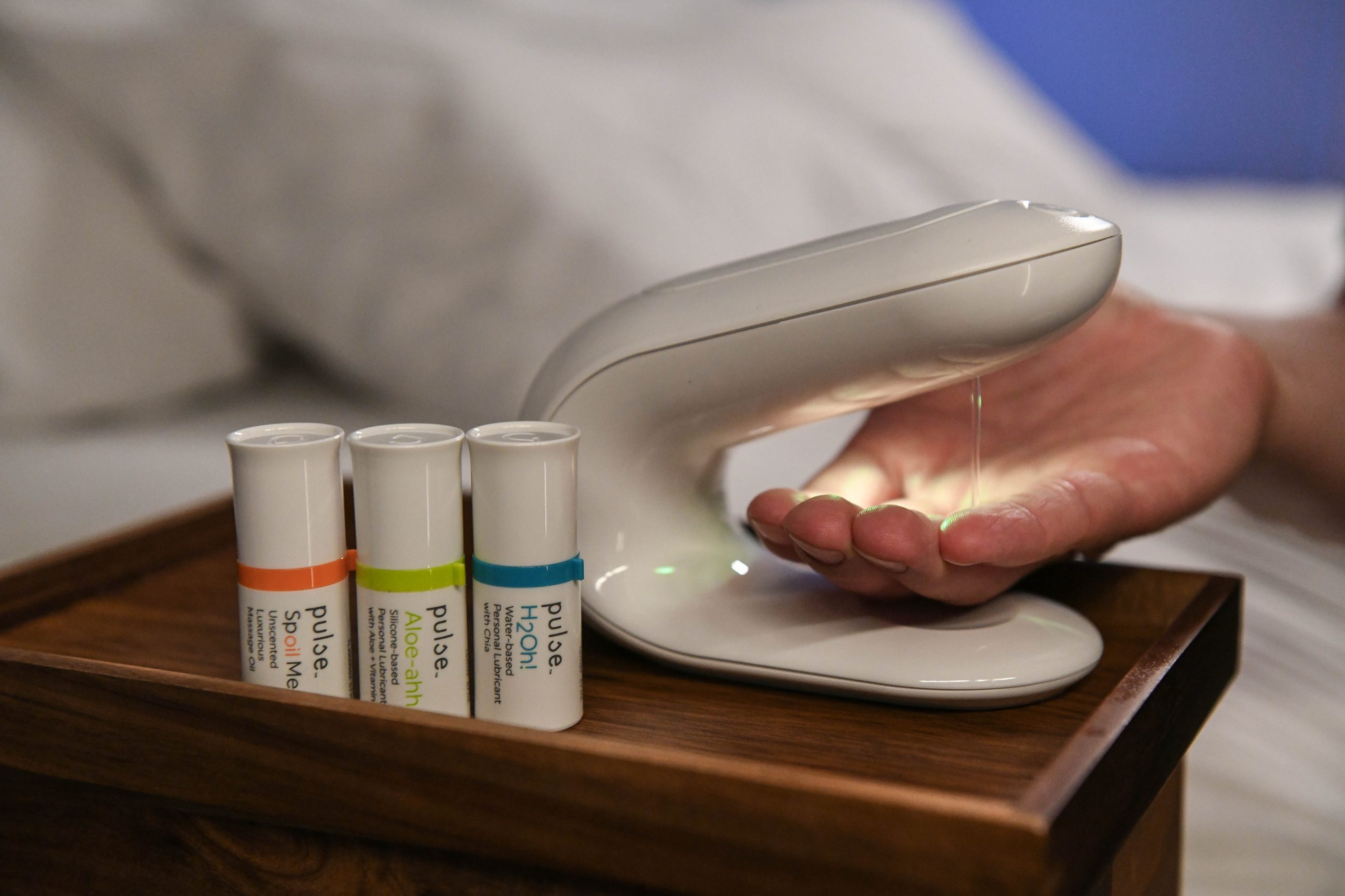 A hand receives liquid from the Pulse Warmer next to massage oil and sexual lubricant pods.